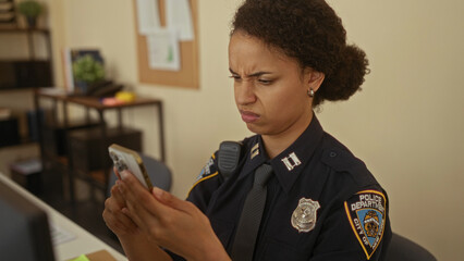 Young woman police officer intently examines smartphone in office setting showcasing dedication and...