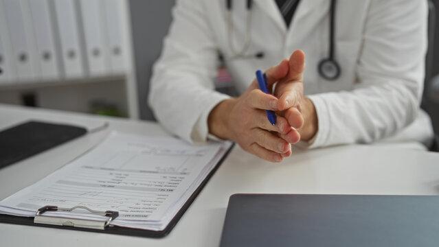 Doctor in clinic wearing white coat sitting at desk with paperwork in front holding pen in clasped hands suggesting consultation in modern medical workplace setting