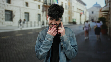 Young man with beard in denim jacket talking on phone outdoors in urban street with blurry...