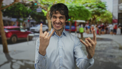 Young man hand making rock horns gesture and smiling on crowded city street during daylight hours;...