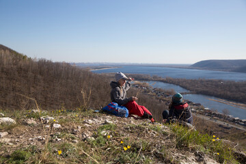 two women  with backpacks relax on a mountain on a hike,  sitting on the grass and enjoy the view  on a sunny spring day.