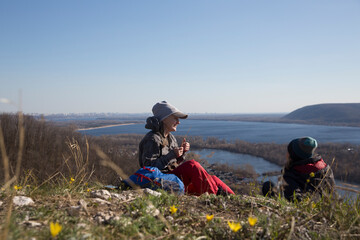 two women  with backpacks relax on a mountain on a hike,  sitting on the grass and enjoy the view  on a sunny spring day.