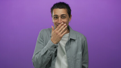 Fototapeta premium Young man covering mouth stands over an isolated purple background showing an expressive reaction with his hand near his face.