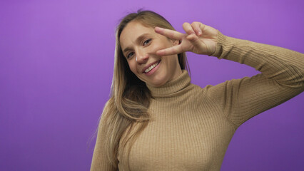 Woman smiling making peace sign on purple background wearing brown turtleneck creating cheerful...
