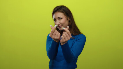 Fototapeta premium Young woman in blue sweater making heart gesture with fingers against a bright yellow background, smiling joyfully in an isolated setting, capturing playful expression.