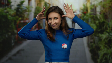 Woman outdoors in city wearing vote badge playfully posing in casual attire with greenery in...