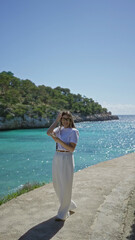 Woman capturing ocean view on smartphone at sunny beach in santanyi, mallorca with vibrant turquoise water and lush greenery under clear blue sky.