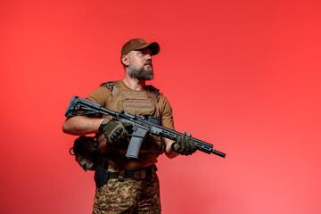 A soldier in military uniform holds a rifle AR 15 in his hands on red background in studio