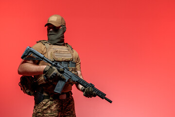 A soldier in military uniform holds a rifle AR 15 in his hands on red background in studio