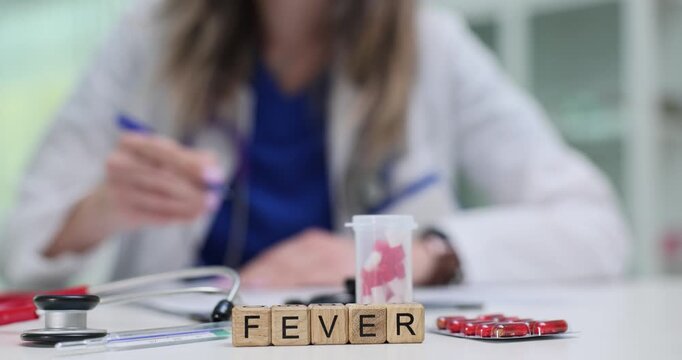 Fever word on wooden cubes stands against doctor by thermometer and red blister pack. Tools suggest temperature check and medication during sickness