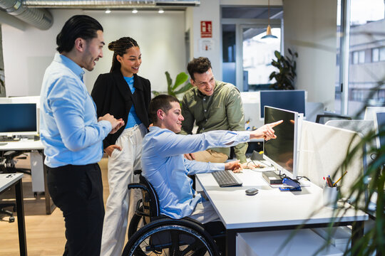Diverse team of young professionals, including a person using a wheelchair, are actively engaged in a computer project, demonstrating teamwork and inclusivity in a modern office environment