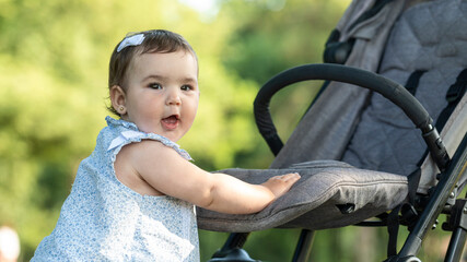 Cute baby girl standing by stroller outdoors in summer park, toddler in light dress with baby...