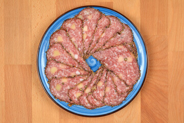 A top-down view shows thinly sliced salami with cheese and peppercorns arranged in a circle on a blue plate, centered on a wooden surface
