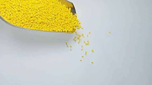 Pouring yellow masterbatch granules with a stainless steel scoop into an isolated glass container with a white background, suitable as a video clip design for industrial plastic manufacturing products