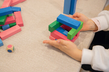 A young child plays indoors during the day, focusing intently while building a structure with colorful wooden blocks, developing fine motor skills, creativity, and early cognitive abilities.