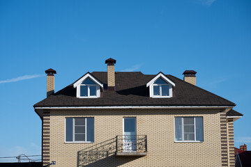 New residential building featuring a pitched roof, multiple chimneys, attic windows, and a small balcony, set against a clear blue sky