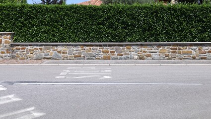 Fence made of low stone wall with high hedge on top. Sidewalk and street with road markings in...