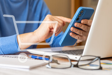 Businesswoman using smartphone for online shopping with a virtual cart icon, while evaluating kpi performance data on laptop with documents, pen and eyeglasses on desk