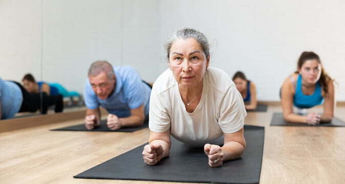 Concentrated elderly woman holding plank pose to strengthen body muscles during warming up before group training in pilates studio. Core exercises for older adults. Active lifestyle concept