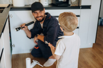 Little boy is helping, holding tool for plumber that is working on the kitchen