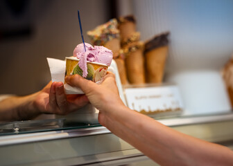 Close-up of a paper cup of purple gelato being handed over a counter, with blurred waffle cones in the background, capturing a friendly ice cream shop moment.