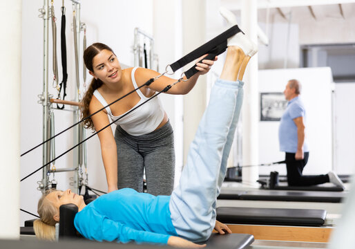 Smiling personal female trainer assisting senior woman exercising on Pilates machine in rehabilitation center