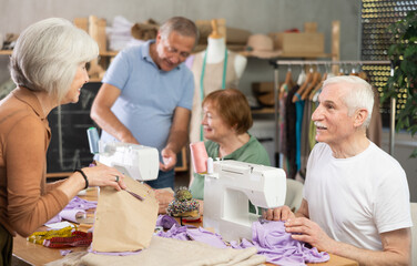 Elderly men and women spending time together sewing in cozy workroom, friendly chatting while using machines, cutting fabric and paper templates during hands-on leisure time