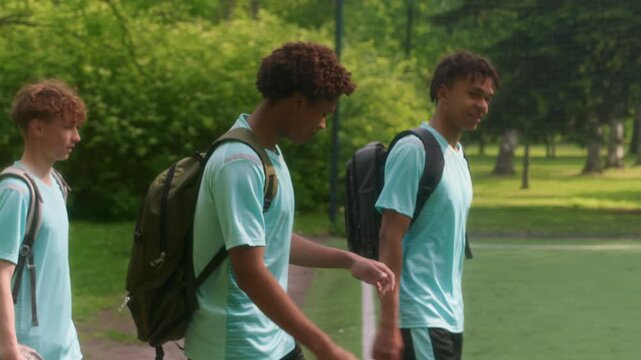 Three teenage soccer players in matching uniforms and backpacks arriving at green outdoor field, ready for practice or match on sunny day in park - Powered by Adobe
