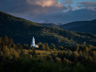 Peaceful white church with tall steeple surrounded by lush green hills and dense forest under a dramatic cloudy sky with golden sunset light perfect for nature and architecture enthusiasts