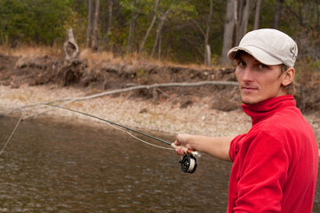A man in a red jacket fishes by the river next to trees and plants