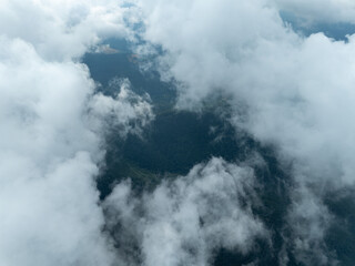 Aerial view of a dense, shadowy forest partially obscured by fluffy white clouds, creating a dreamy, ethereal landscape, Oderen, Grand Est, France.