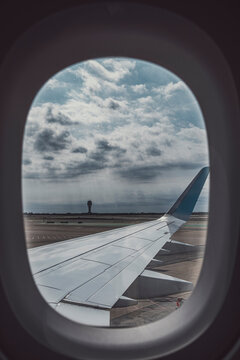 Airplane wing seen through window while aircraft is grounded, cloudy skies and distant control tower visible, representing anticipation of air travel and mobility.