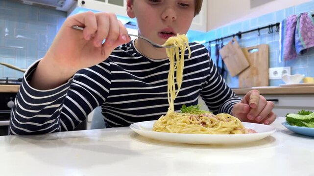 Caucasian boy enjoy eating with spaghetti carbonara with cheese and bacon on a plate in lunch time at the kitchen.