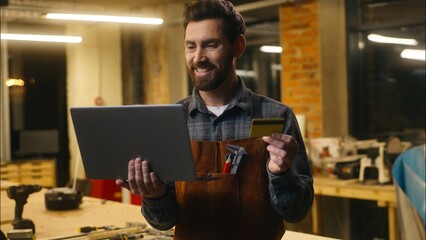 Cheerful Caucasian man wood master hold credit card typing laptop in workshop male client carpenter builder entering security number guy workman paying finance shop online craftsman Internet banking