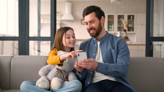 Smiling Caucasian child little girl dad father man touching screen mobile phone smartphone speaking family at home e-commerce glad kid daughter online shopping together parent male Internet cyberspace - Powered by Adobe