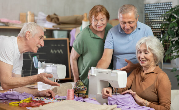 Enthusiastic older adults, women and men, gathering around sewing machine in training workshop while female seamstress explaining stitching techniques and fabric positioning, sharing professional tips - Powered by Adobe