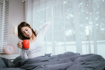 A young woman relaxes in bed, holding a red mug and smartphone, smiling while browsing or chatting,...