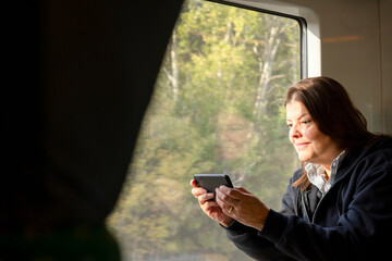 Woman using smartphone on a train, enjoying the scenic view through the window. Leisure or remote work while traveling by rail on a sunny day.