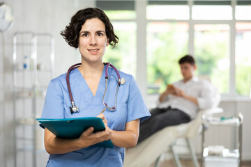 Friendly young female medical person writing at folder standing in treatment room of medical center