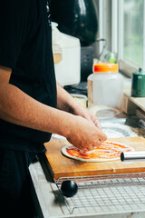 Close-up of hands adding fresh basil leaves to a pizza topped with ham and tomato sauce on raw...