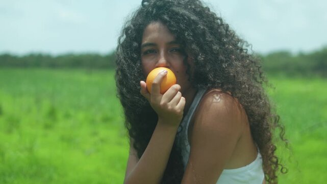 Young woman with voluminous Type 3C curls and warm brown skin holds a peach-like fruit close to her mouth, staring at the camera in a vivid green field under bright midday light.