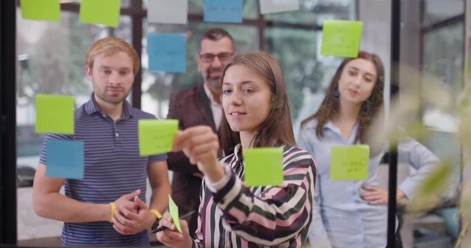 Team of caucasian business people standing near glass wall with sticky notes. Scrum methodology with colorful stickers. Work colleagues discussing project ideas.