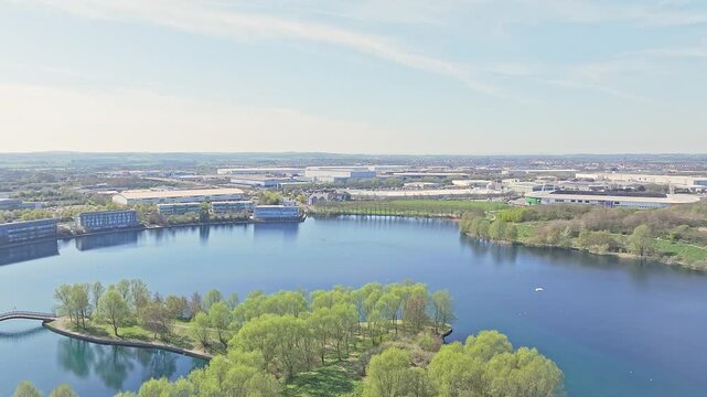 Drone ascending over Doncaster Lakeside in South Yorkshire, England, showing a vibrant green island surrounded by calm waters, with office parks, commercial buildings, and distant horizon in view.