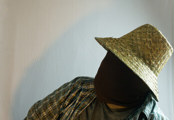 Close-up of masked male farmer wearing straw hat, face fully covered in brown cloth, white...