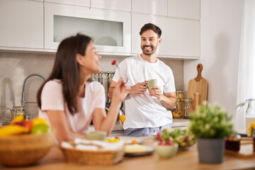 A couple is seen in their bright kitchen, smiling and chatting as they enjoy a light breakfast with drinks and fresh produce on the counter, creating a cheerful atmosphere.