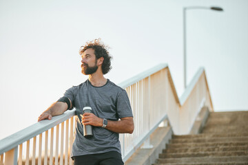 A man stands on the steps after a workout, gazing thoughtfully into the distance while holding a...