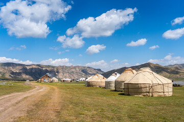 A collection of traditional yurts in a wide mountain valley, with a dirt road leading towards distant hills under a vibrant blue sky