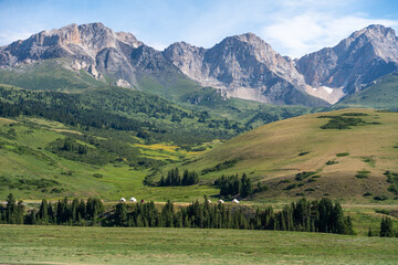 A picturesque mountain valley featuring a line of yurts nestled among green forests and rolling hills, with rugged peaks in the background