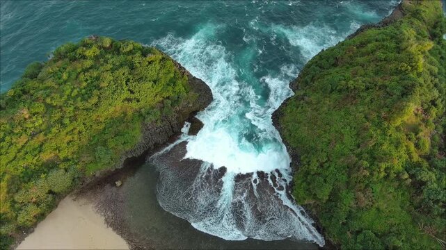The aerial view shows waves continuously crashing into the gap between two cliffs covered with green trees in the afternoon.