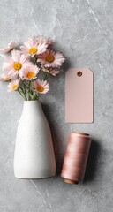Pastel pink flowers in a white vase, a tag, and thread on a gray stone surface